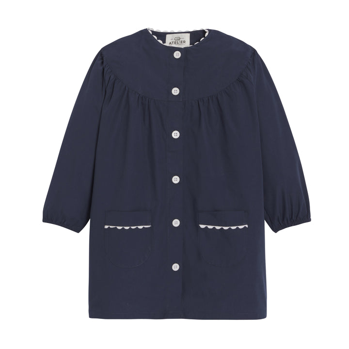 Fille wearing a navy blue school apron from the Jeanne collection by My Store, featuring a front pocket and adjustable straps.